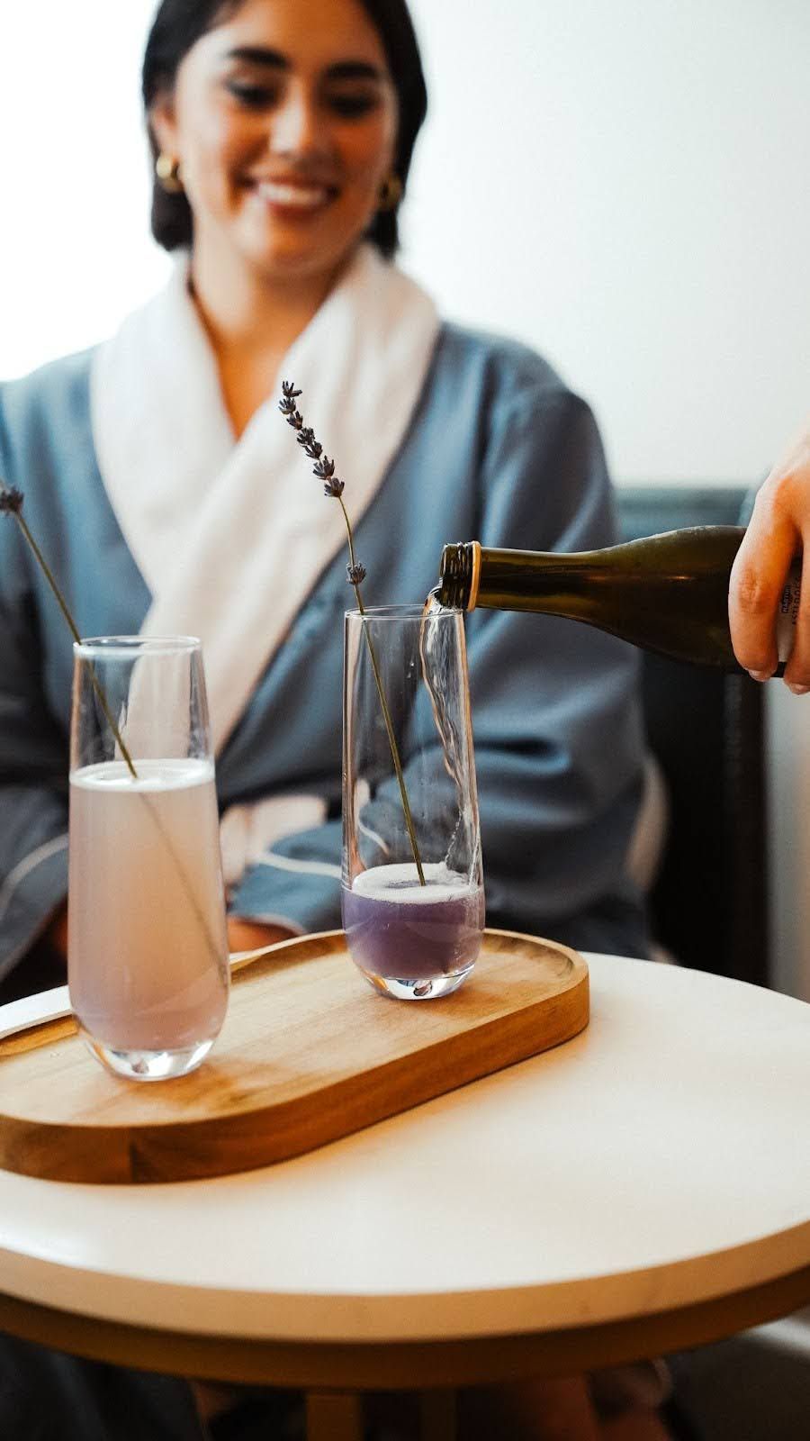 Woman in robe smiles as champagne is poured into lavender-infused glasses on a wooden tray.