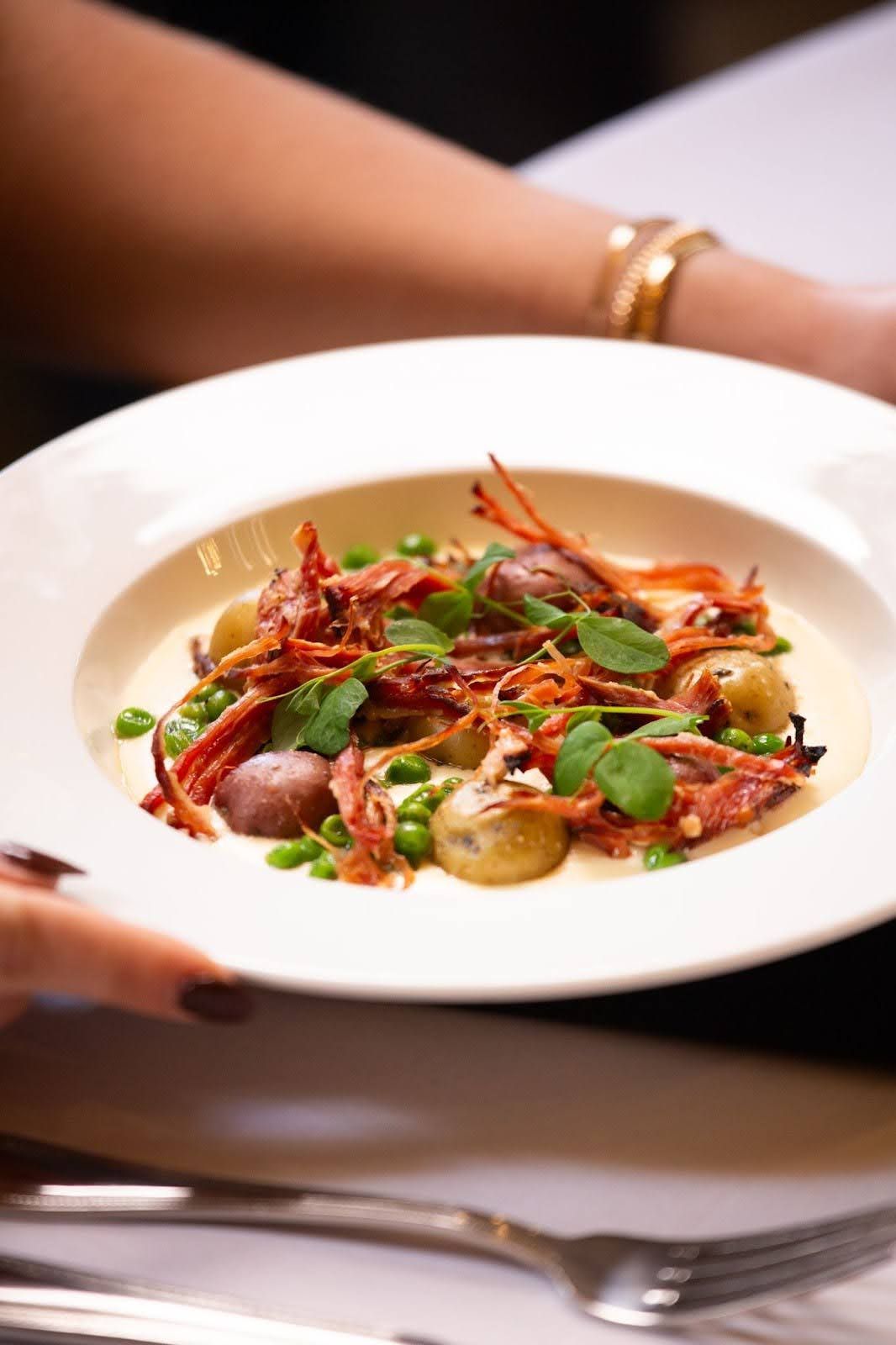 Person holding a white plate with a creamy dish of potatoes, peas, and crispy meat.
