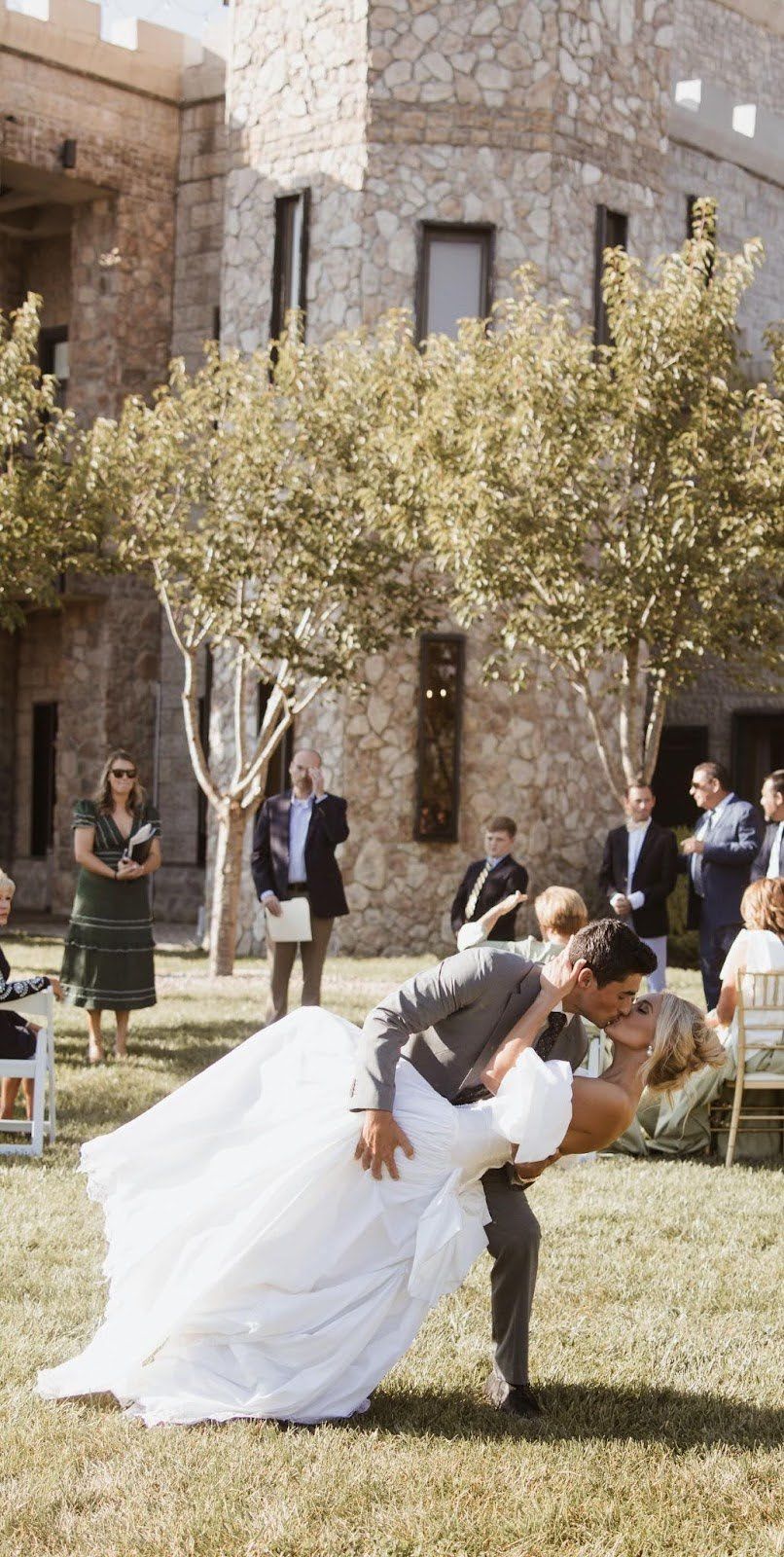 Bride and groom share a kiss, bending backward on grass. Guests and castle in the background.