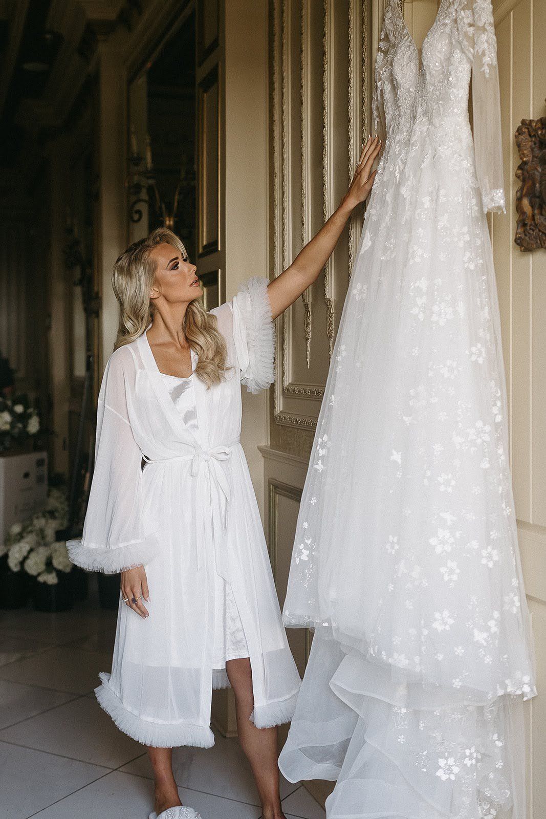 Blonde woman in a white robe admires her wedding dress hanging on a wall in a luxurious room.
