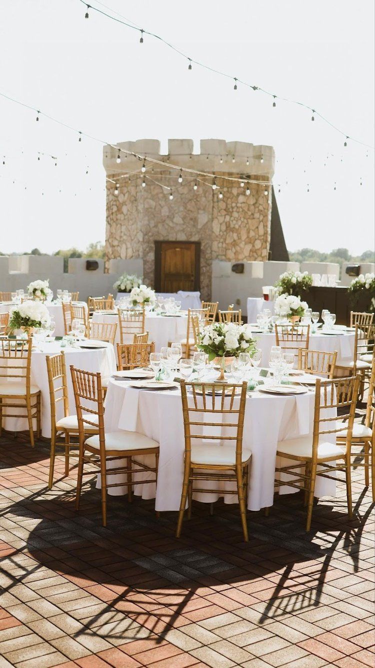 Outdoor wedding reception with round tables set with white linens and gold chairs, against a castle backdrop.