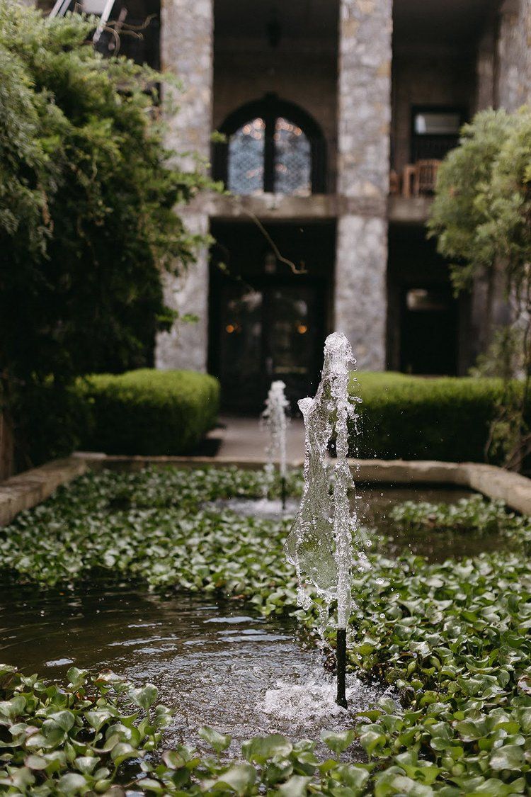 Fountain sprays water in a lily-filled pond in front of a stone building with arched windows.