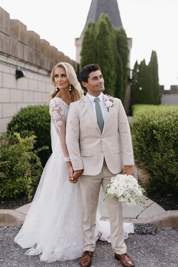 Bride and groom holding hands on wedding day in front of a castle, the groom looking to the side.