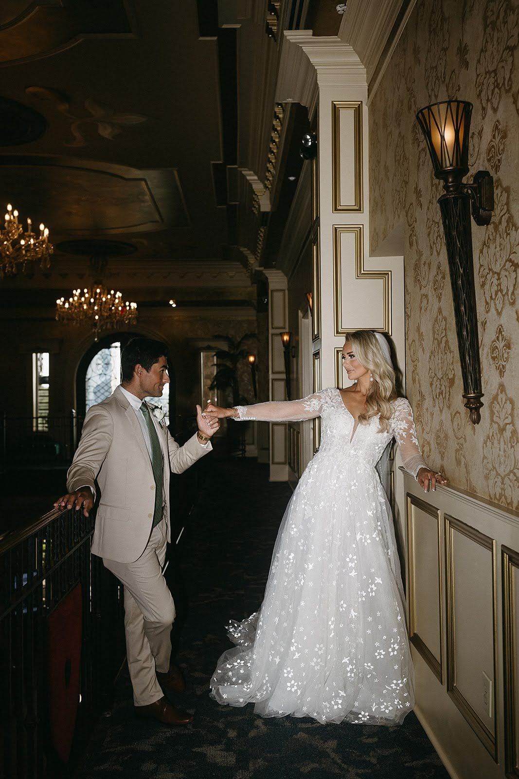 Bride and groom holding hands on a balcony. She wears a white gown, he wears a beige suit. Elegant interior.