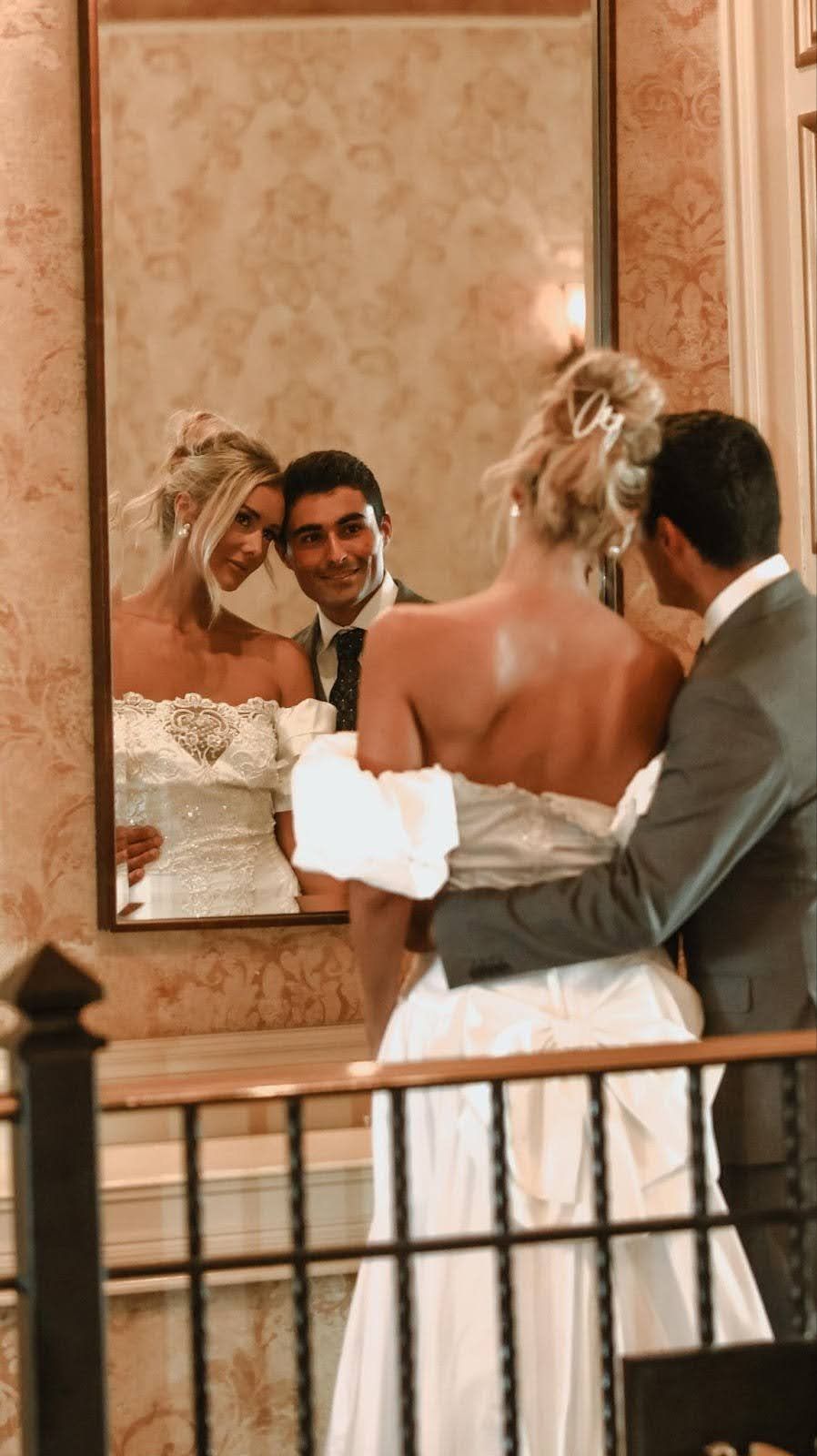 Bride and groom posing, arms around each other, reflected in a mirror. Elegant wedding attire, interior setting.