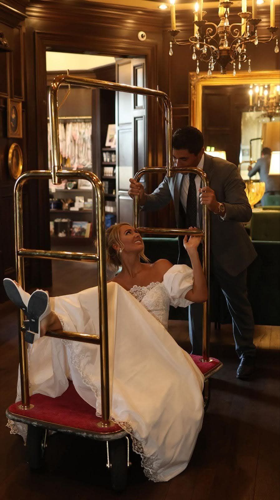 Bride in gown sits on a luggage cart as groom pushes it, indoors.