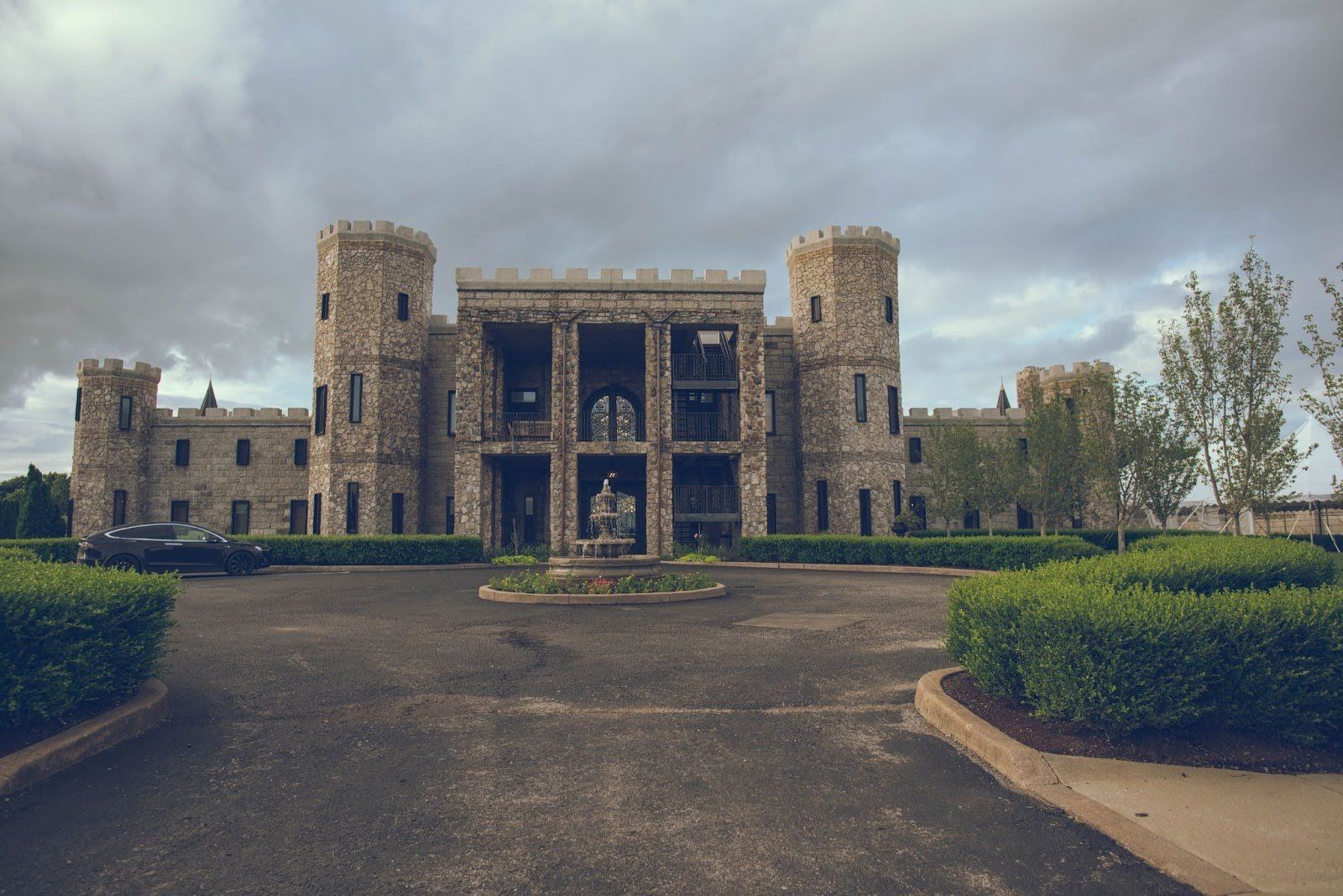 Castle with stone facade, turrets, and central open section. Gray sky overhead, with a fountain and shrubs in front.
