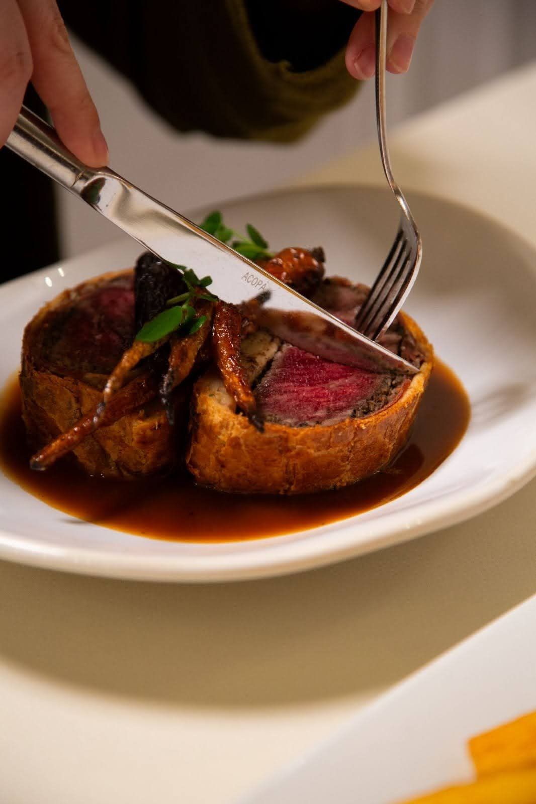 A person cutting a wellington dish; a browned pastry crust with a rare, red meat center; sauce.