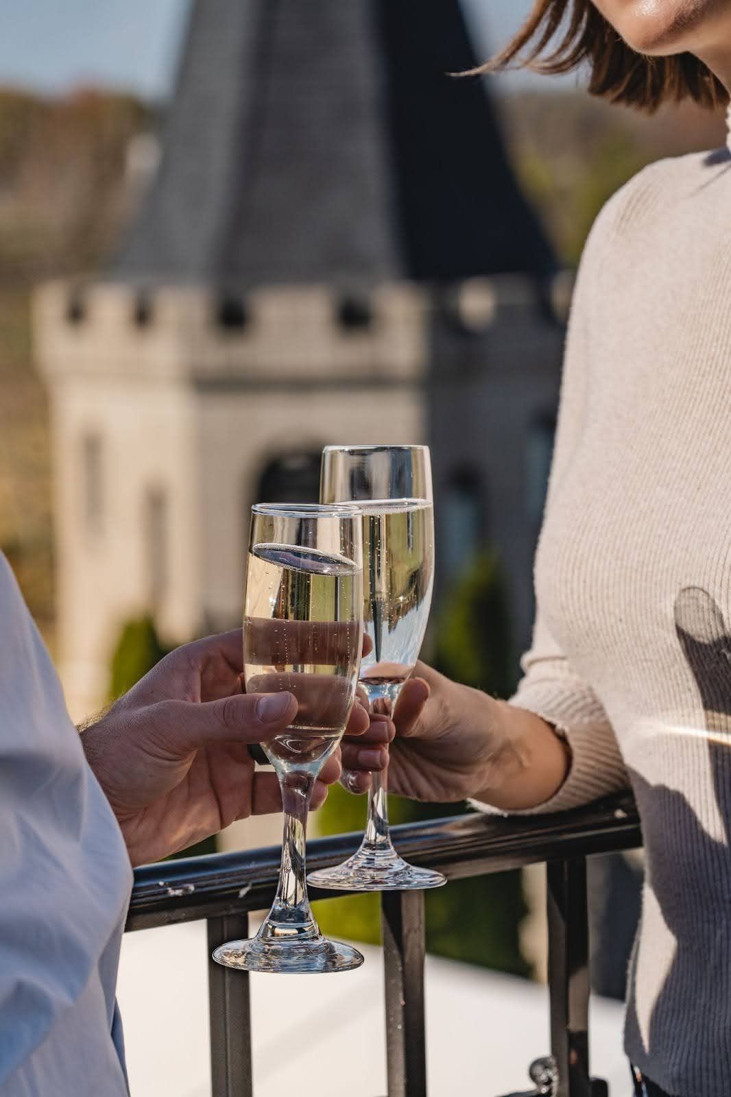 Two people toast champagne flutes on a balcony, castle tower in the background.