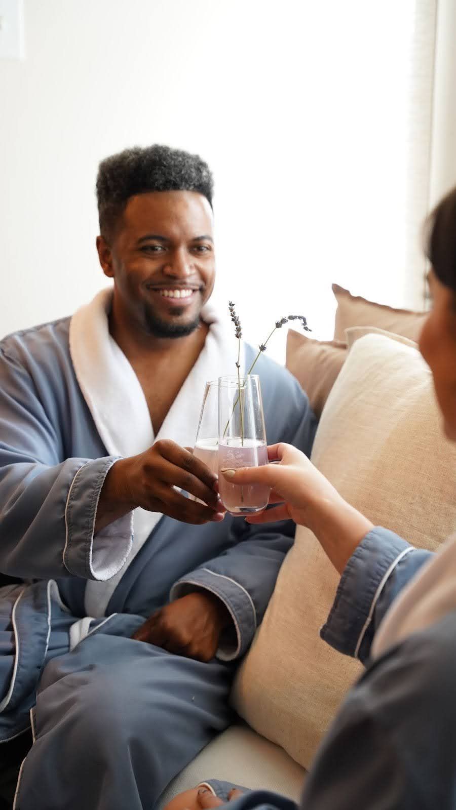 Man in robe clinks glasses with a person, smiling. Indoors, light blue robe, beige pillow.