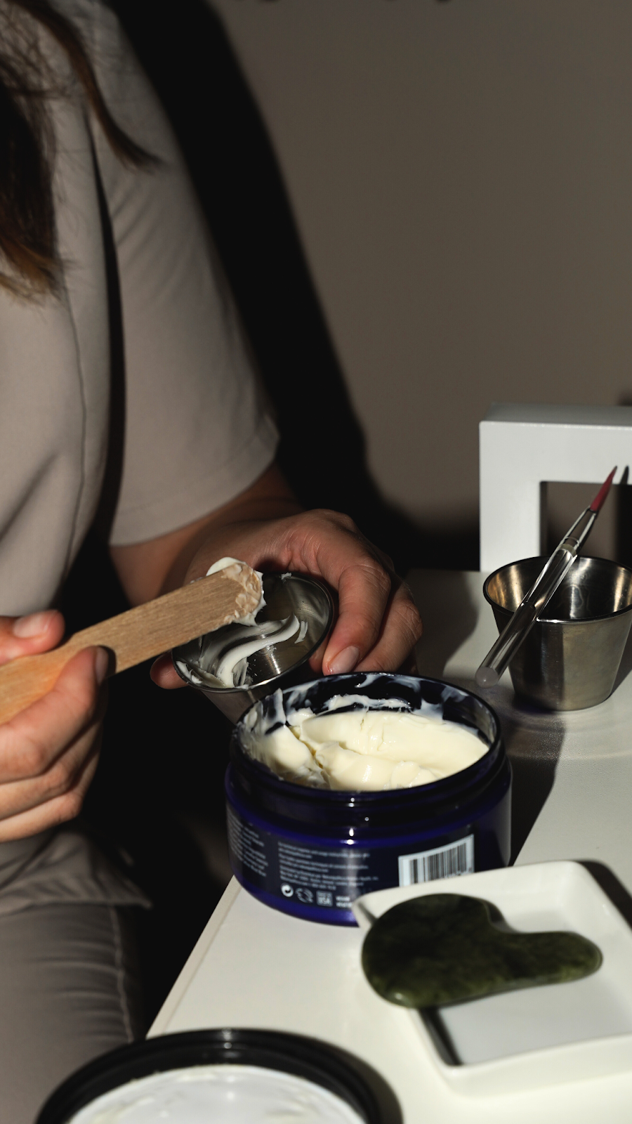 Person scoops cream from a blue jar with a wooden stick, preparing skincare on a white surface.