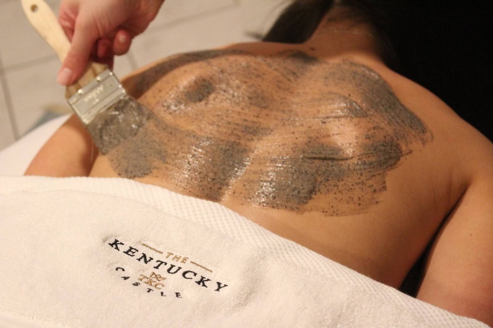 Person receiving a back treatment at a spa. A hand uses a brush to apply grey product on the back.