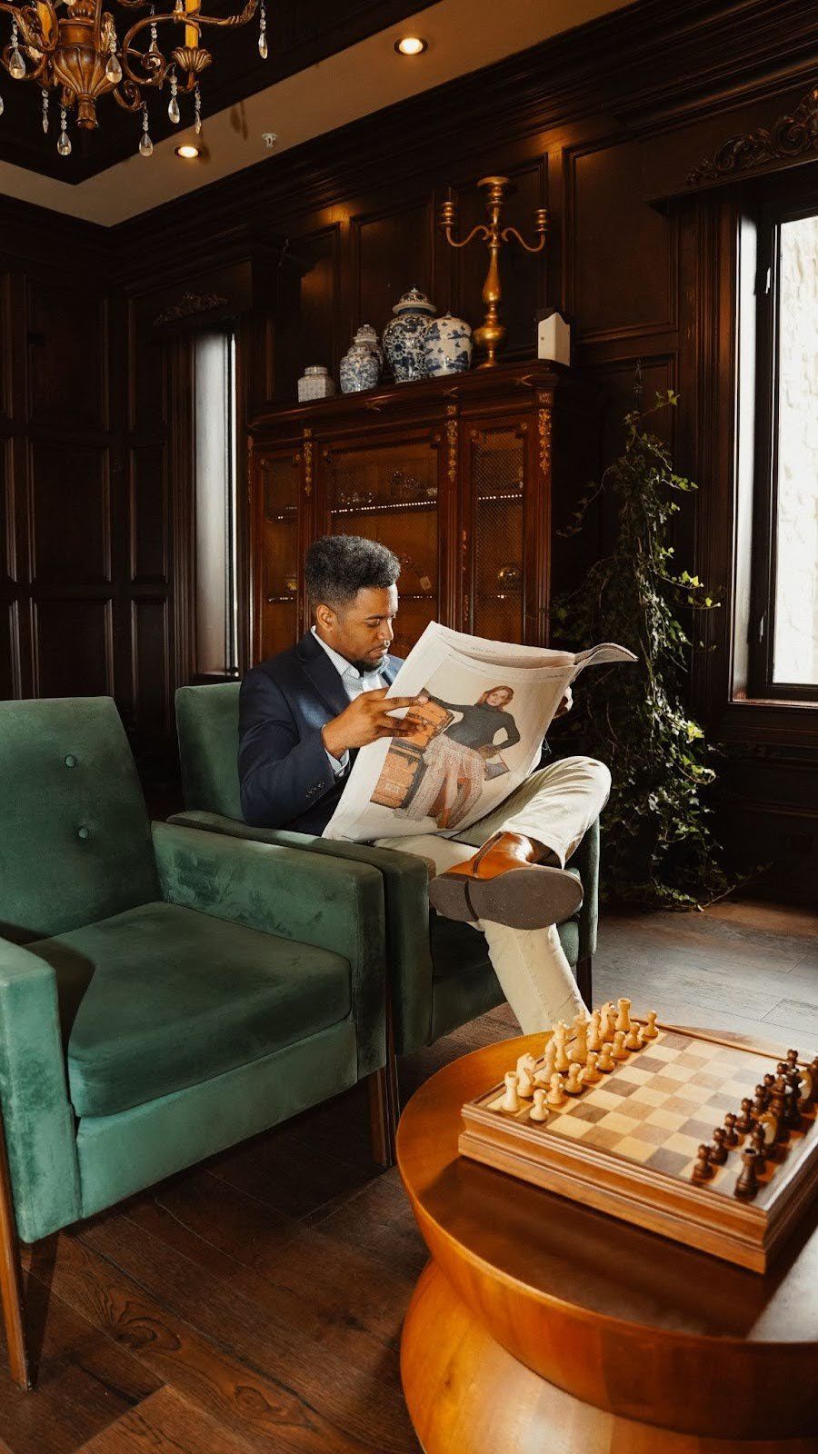 Man in a suit reads newspaper in a wood-paneled room, seated in a green armchair near a chess board.
