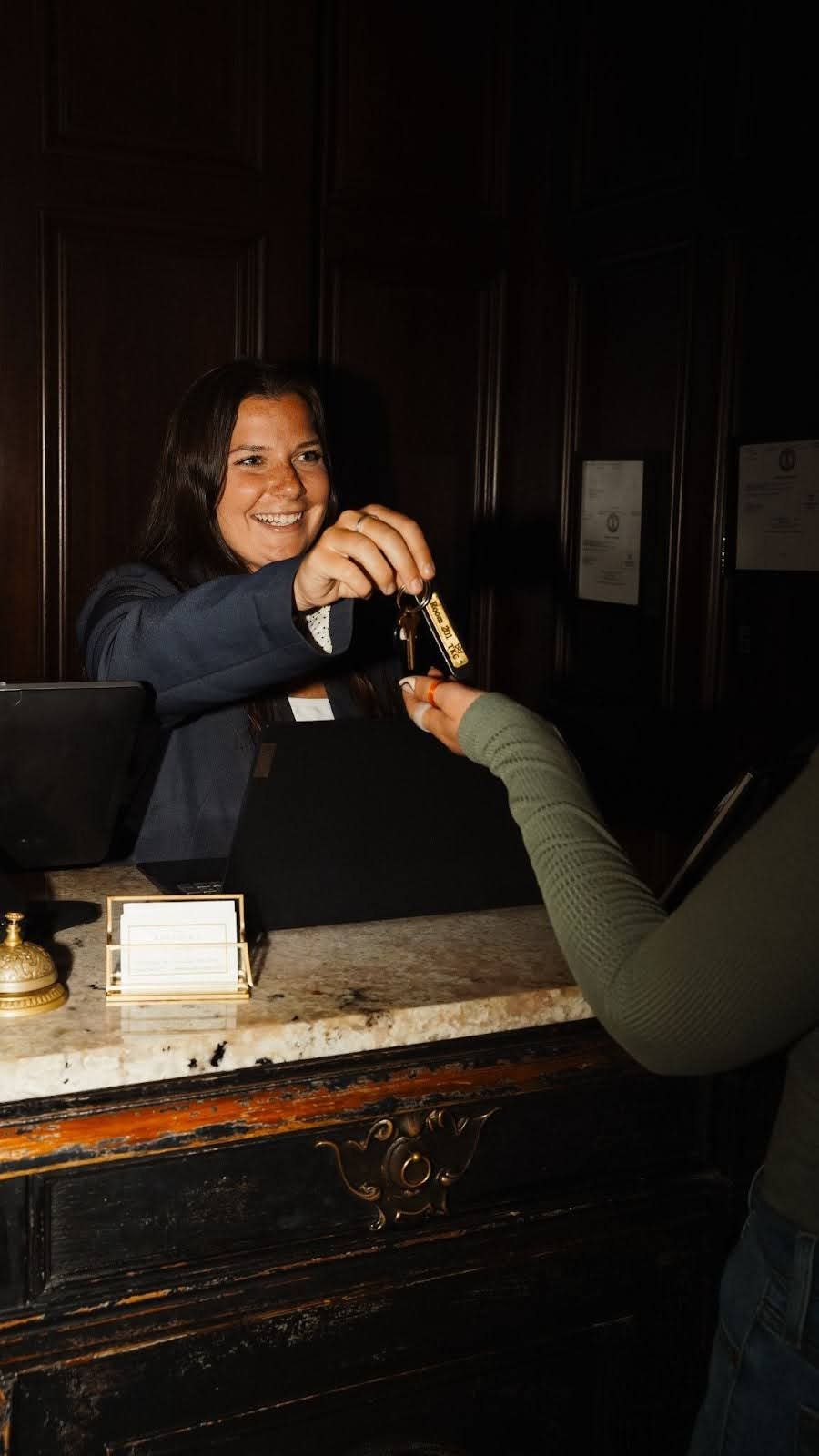Hotel clerk handing keys to a guest at a dark wooden reception desk.
