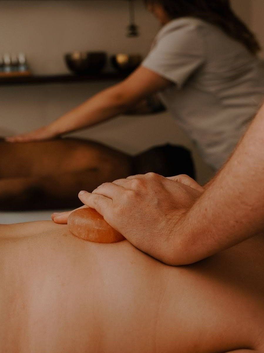 Person receiving back massage with another in background. Spa setting with bowls and shelves.