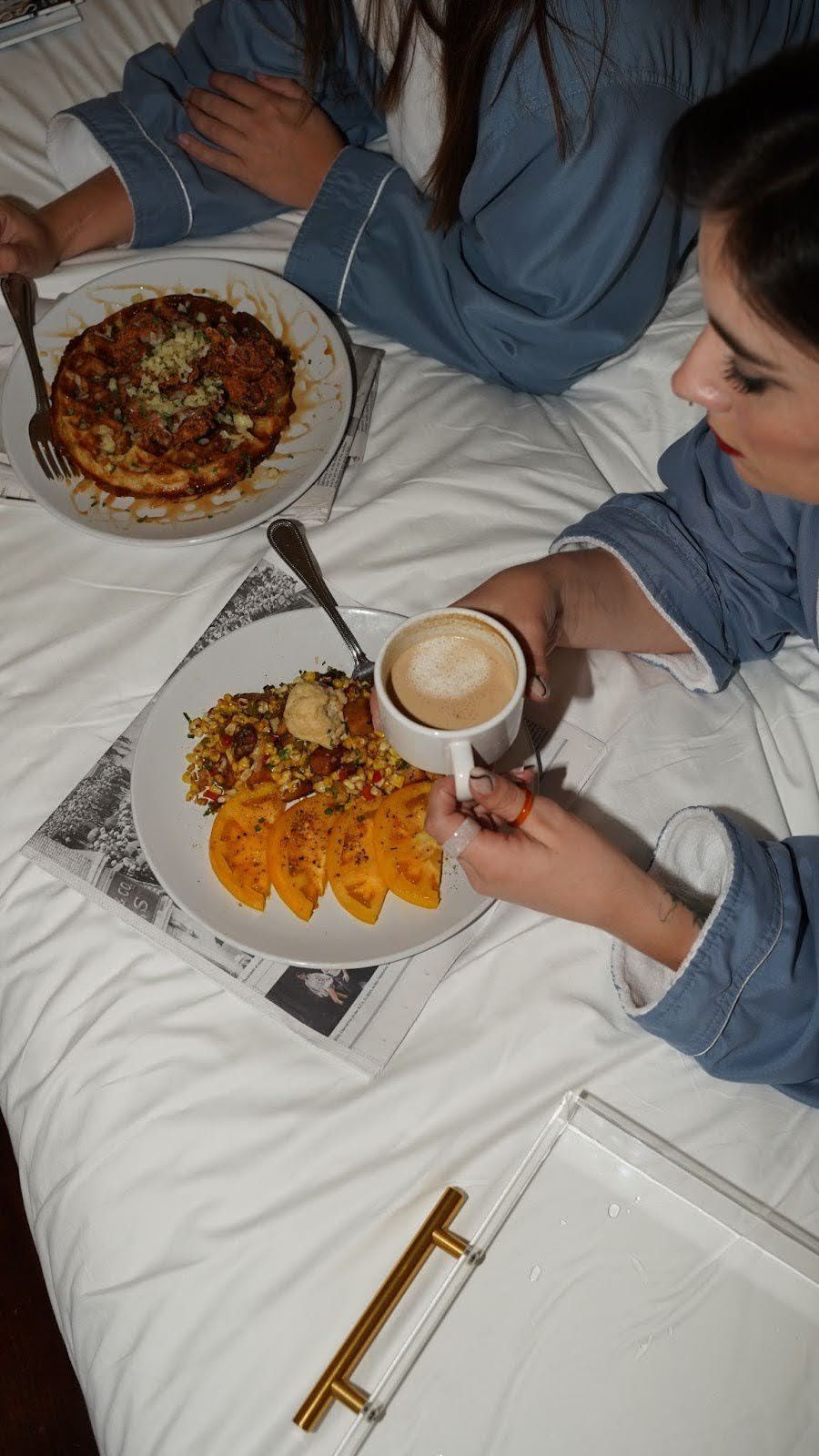 Two women in robes eating breakfast in bed: pancakes, waffles, coffee.