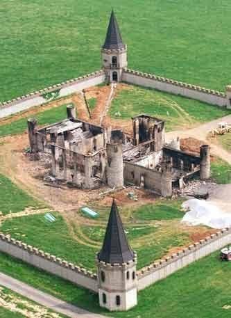 Burnt-out castle ruin within a fortified green wall. Two tall towers flank a damaged main building on grassy land.