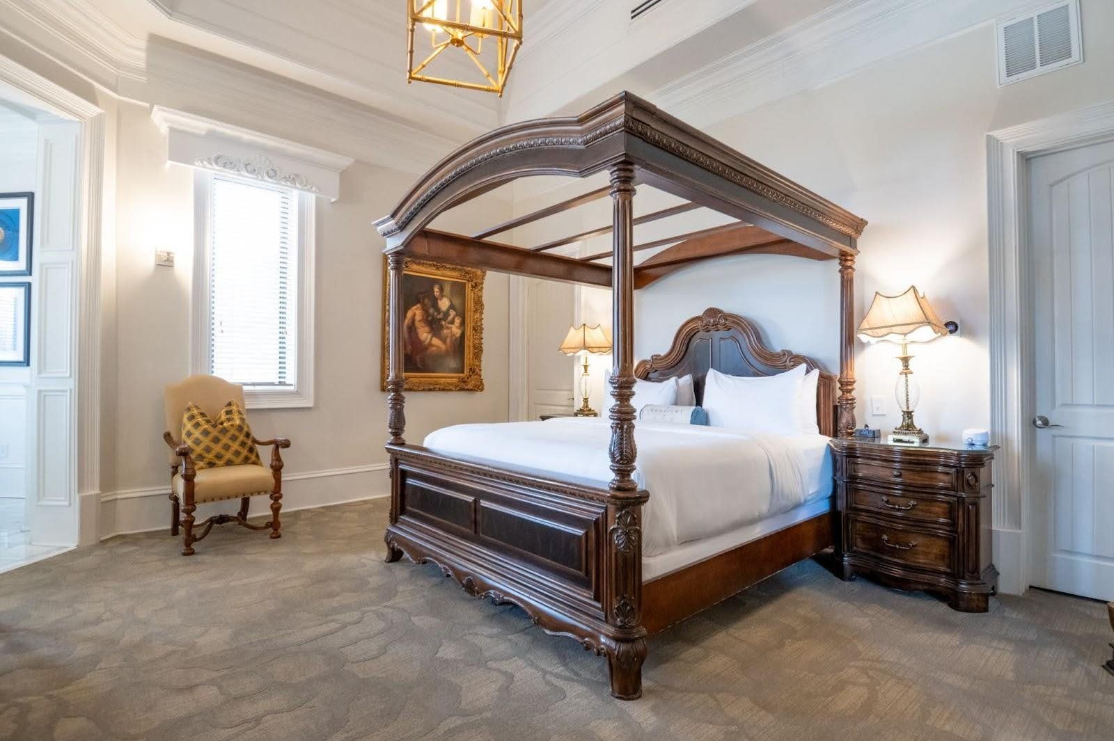 Bedroom with four-poster bed, nightstand, chair, window, and artwork.