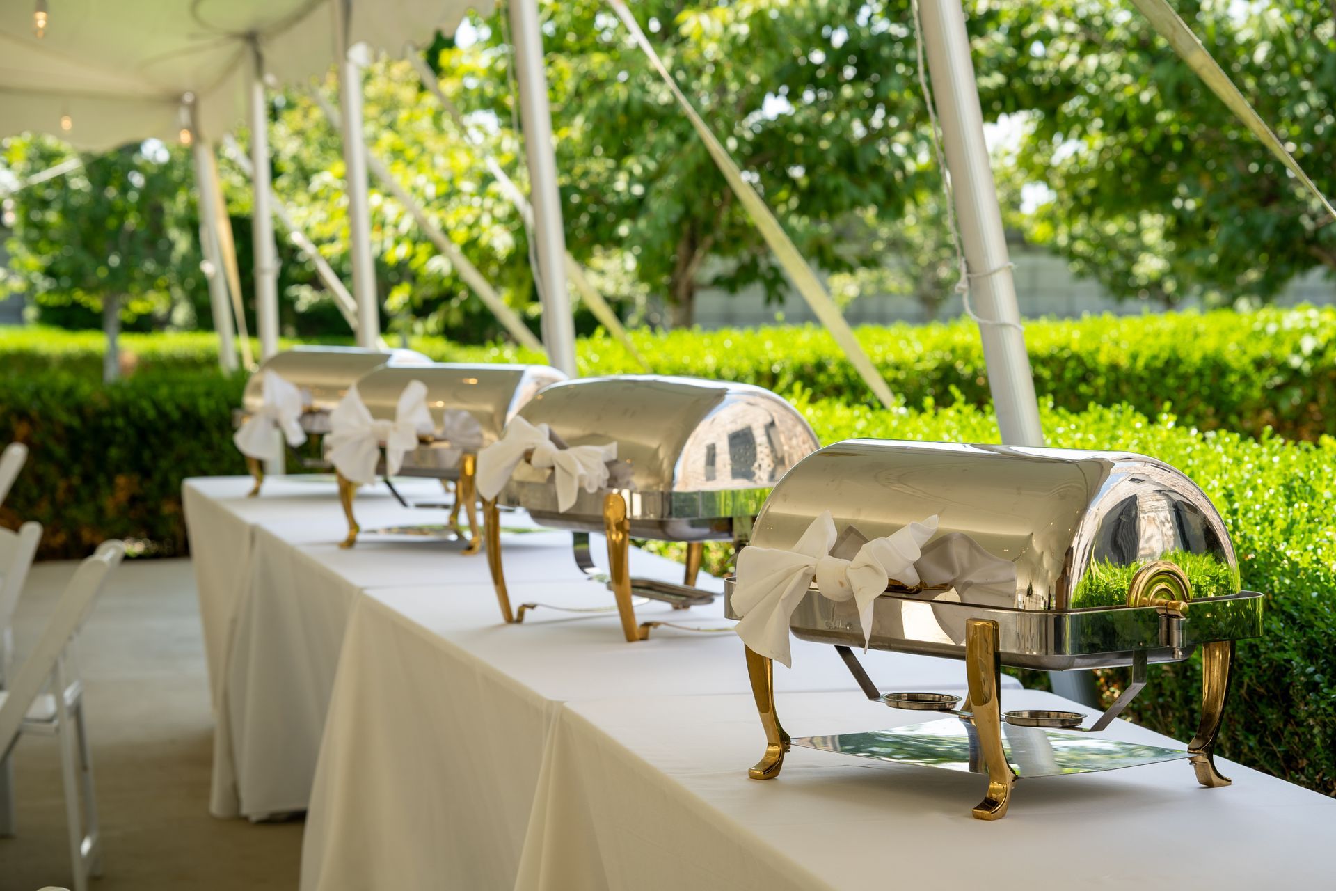 Buffet table with silver chafing dishes, white tablecloth, and greenery in background.