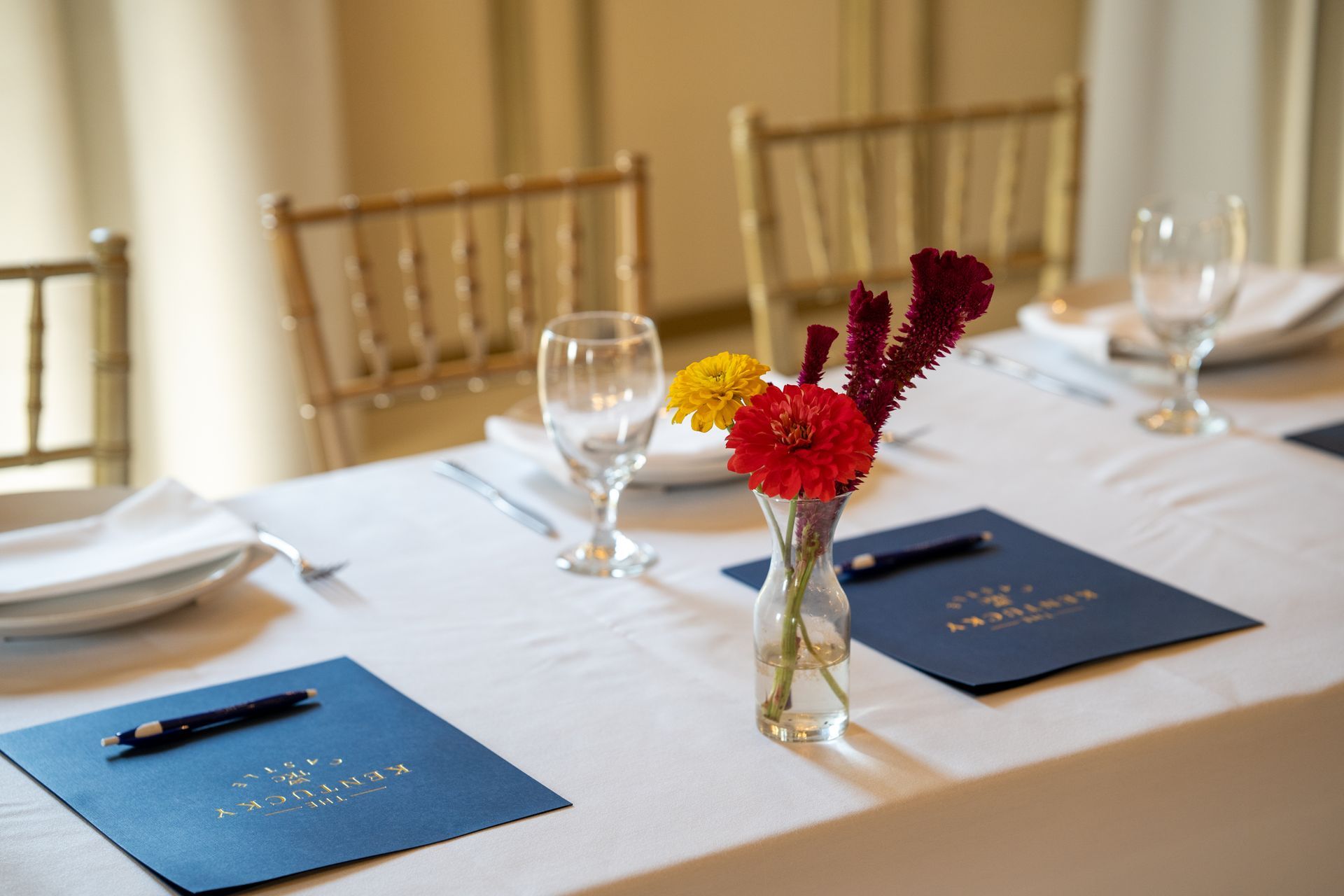 Table setting with flowers, menus, and glasses; gold chairs in a well-lit room.