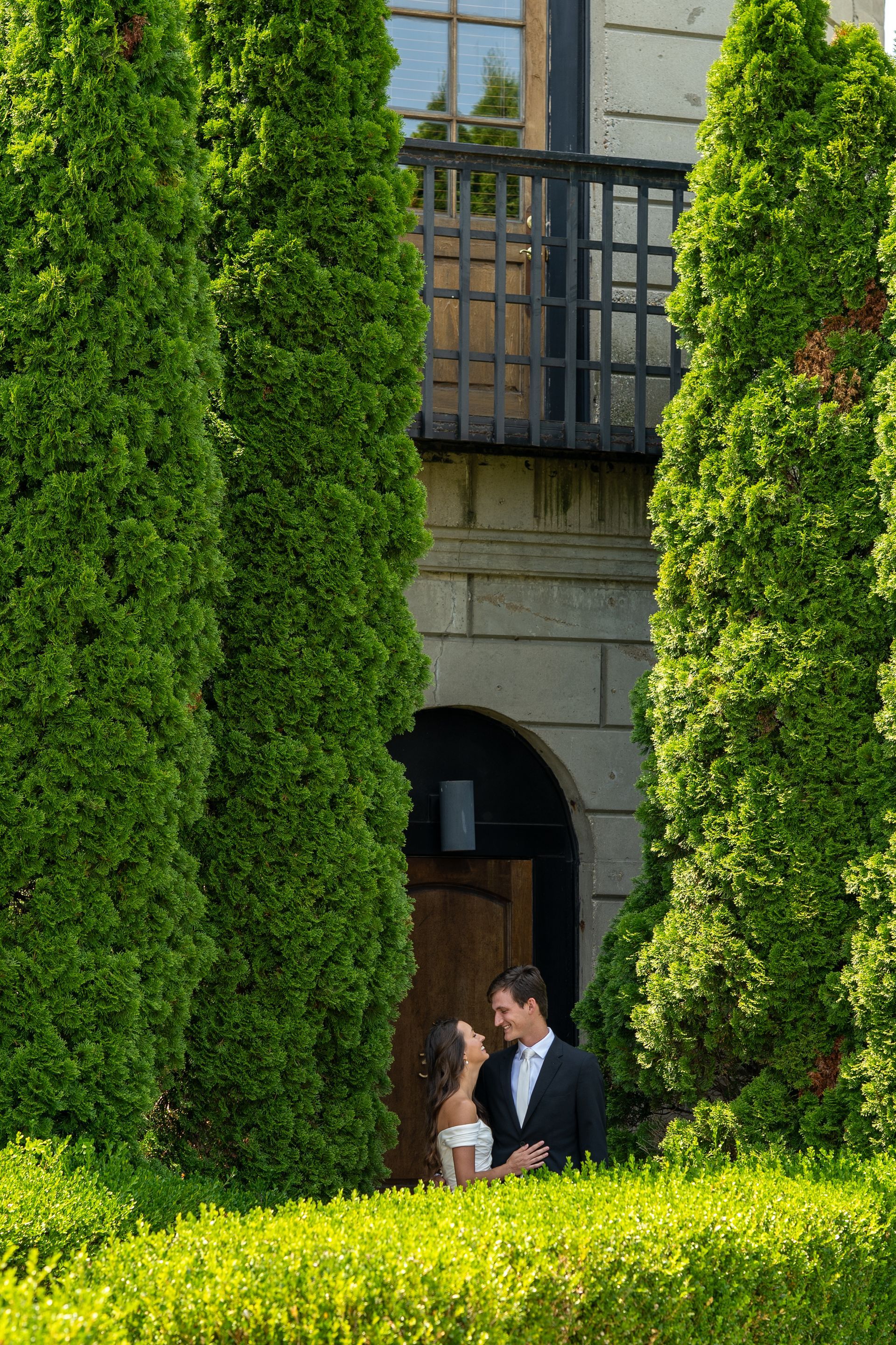 Couple in wedding attire, framed by green trees, facing each other in front of a stone building.