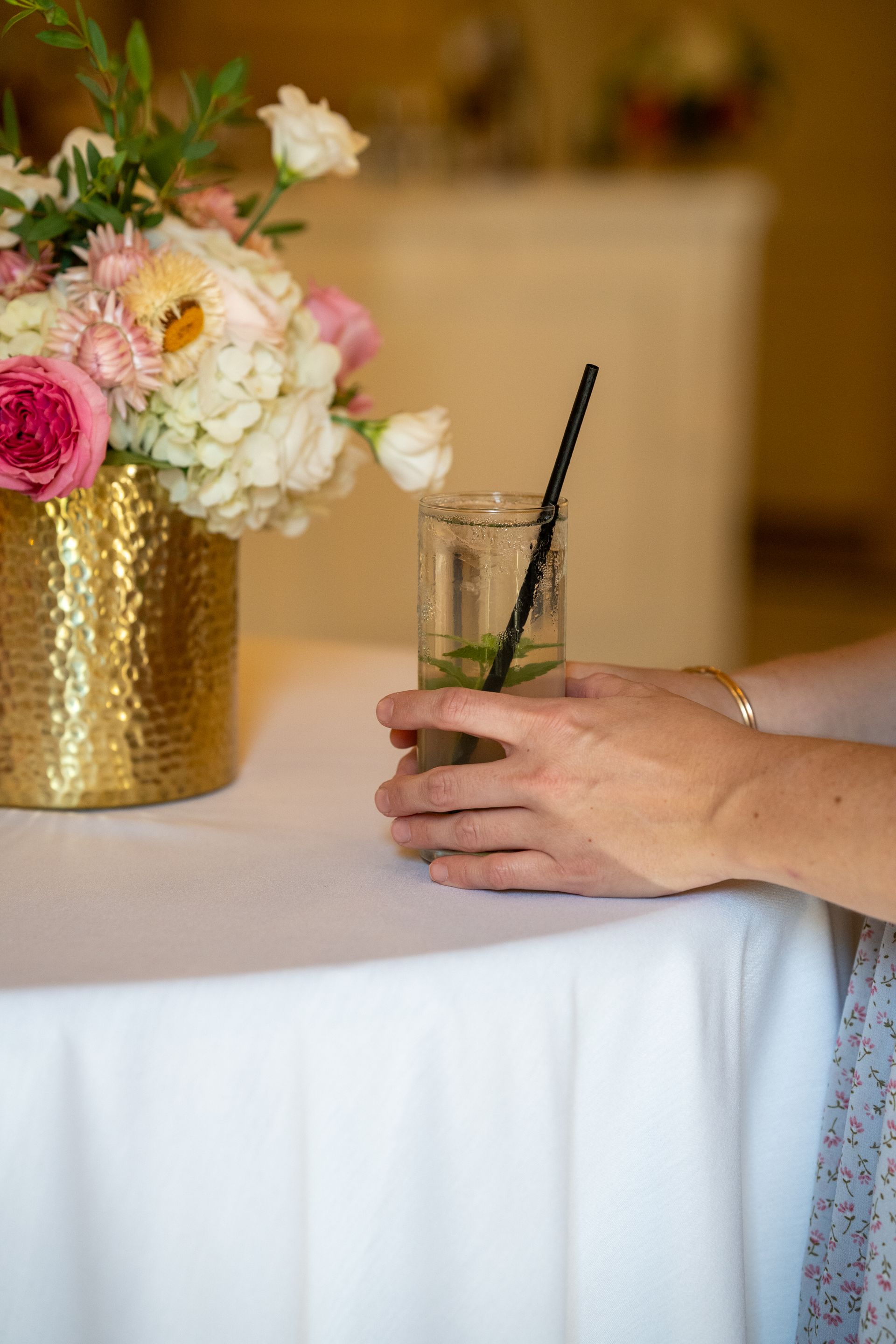 Person holding a cocktail with a black straw, near a vase of flowers on a white tablecloth.