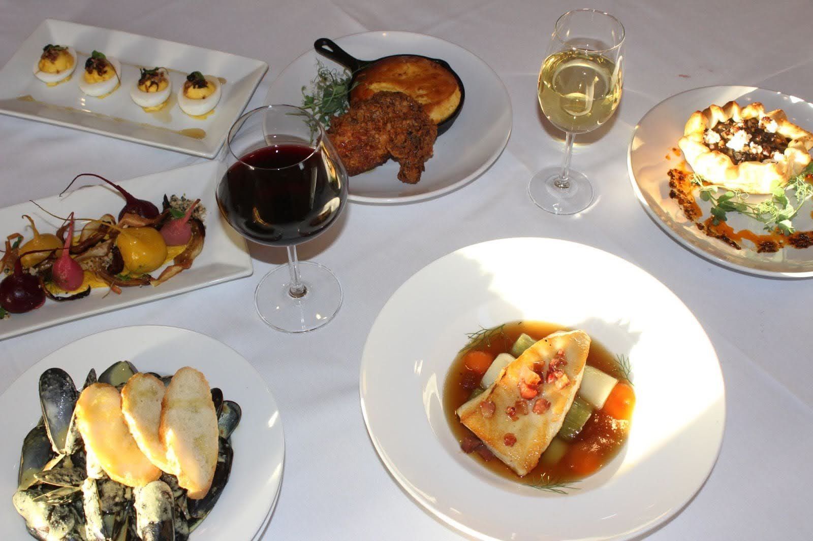 Multiple dishes on a white tablecloth, including deviled eggs, fried chicken, fish, and mussels, with wine glasses.