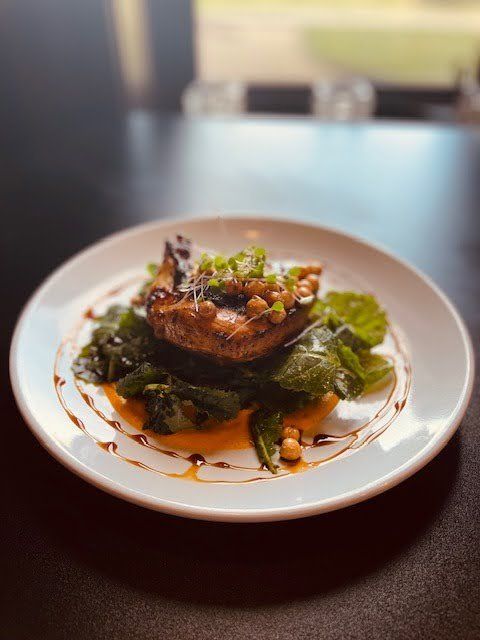Plate of seared meat on a bed of greens, drizzled with sauce and garnished with herbs on a white plate.