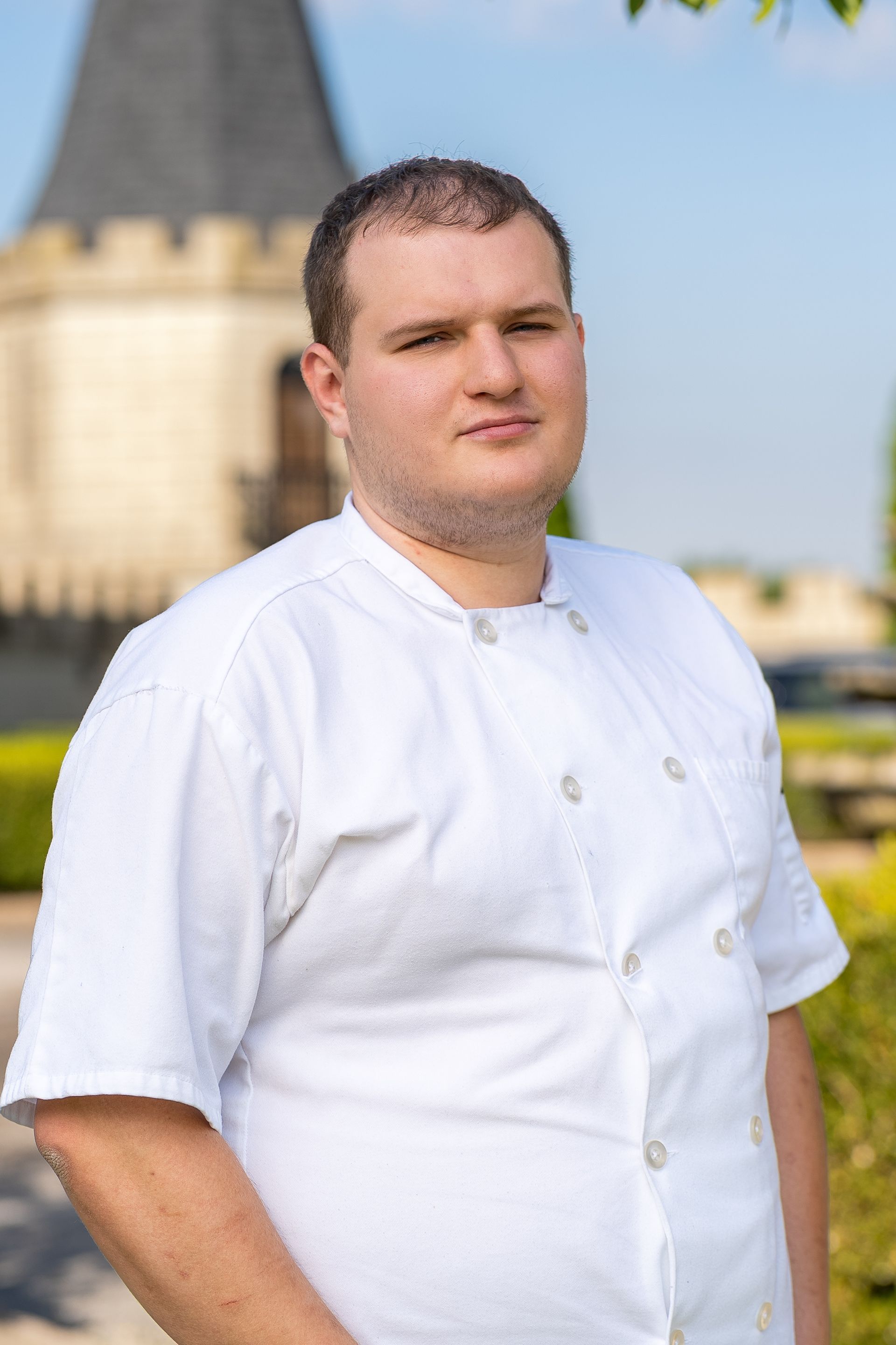 Chef in black uniform, glasses, smiling in a kitchen, ovens in background.