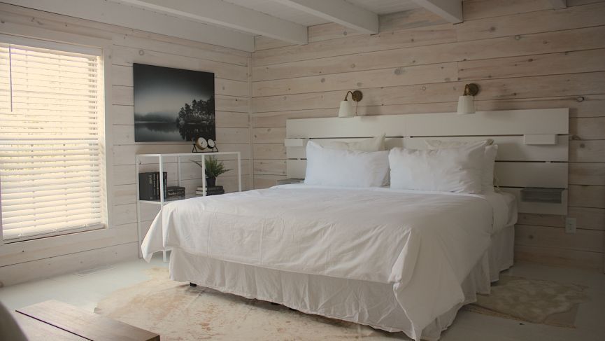 White bedroom with a bed, window with blinds, artwork, and a small shelf.