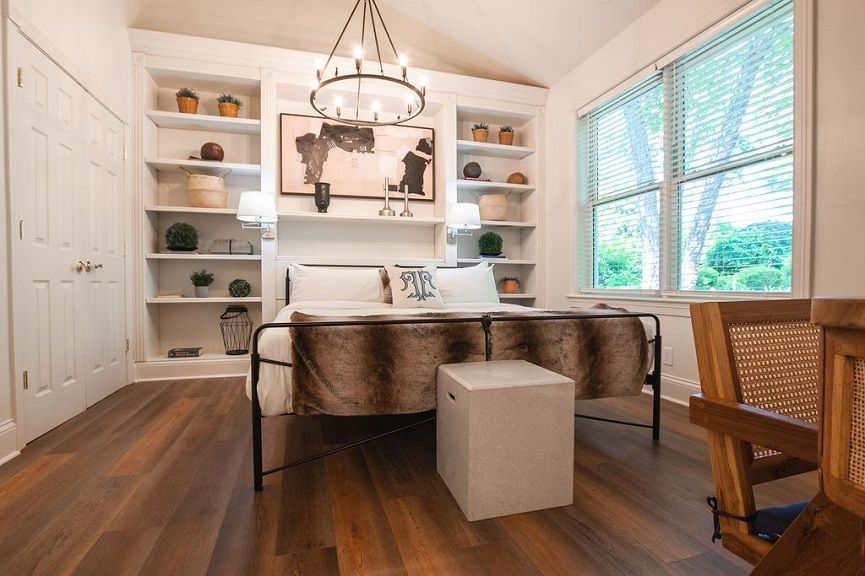 Bedroom with built-in shelves, bed with fur throw, painting, white cube, and window with green view.