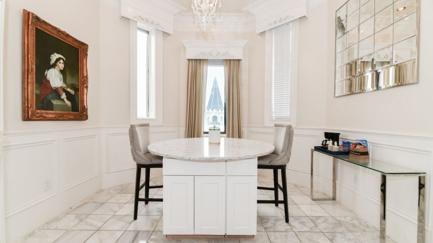 Elegant white room with a round table, two chairs, a painting, and a small bar; marble floor.