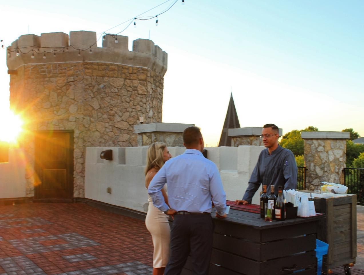 People at outdoor bar on rooftop with castle turret at sunset.