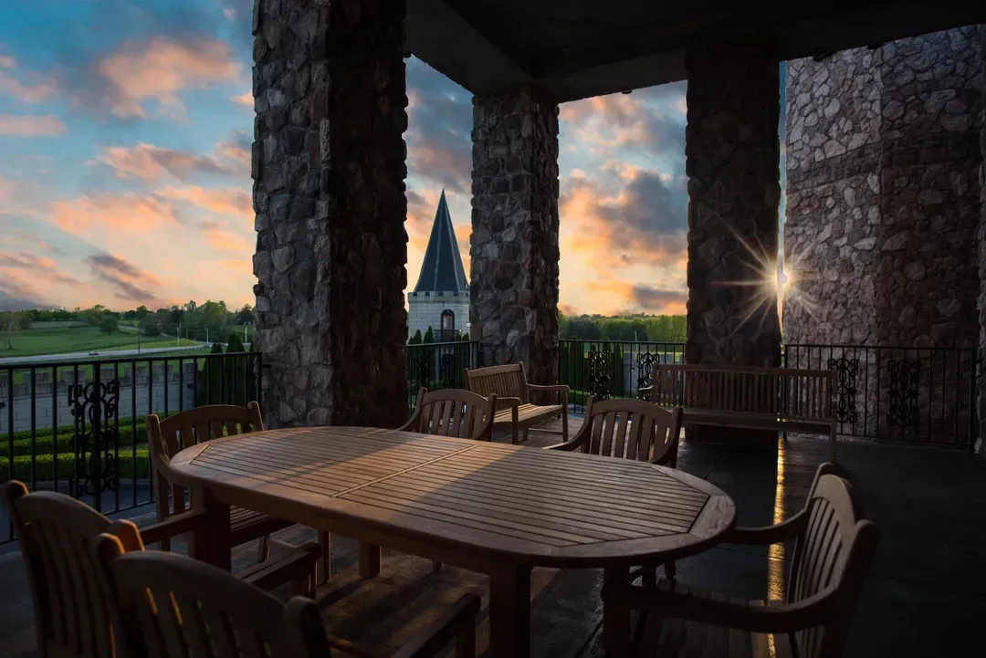 Patio with wooden furniture, stone columns, and a sunset view of a building with towers.