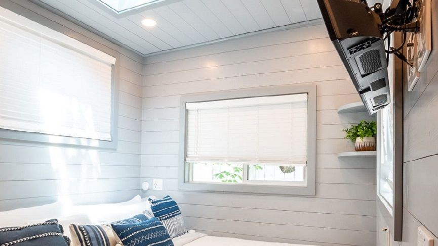Cozy bedroom with white blinds, shiplap walls, pillows, and a mounted TV.