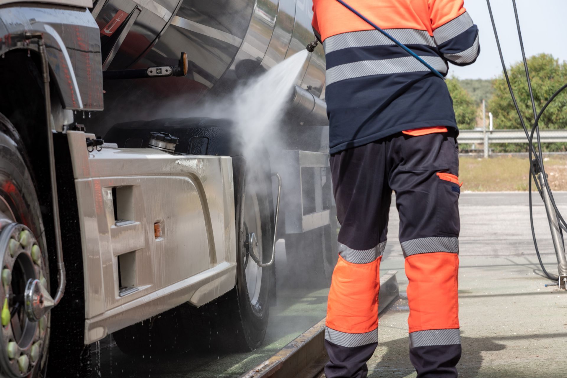 Person in orange workwear washing a truck with a water hose at an outdoor car wash.