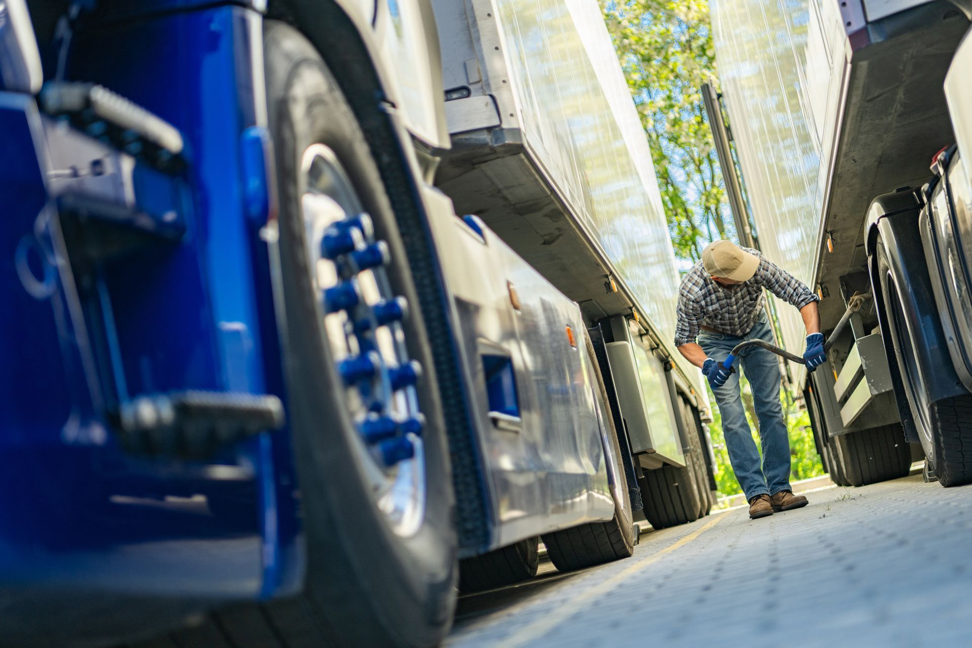 Man in jeans and plaid shirt works on trucks outdoors; blue semi-truck wheel in foreground.