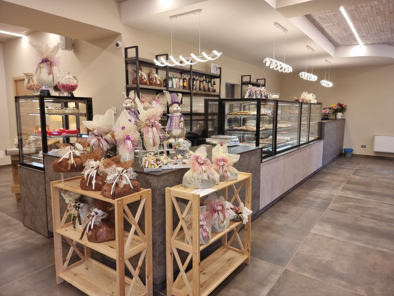 Interno di una panetteria con vetrine di dolci, scaffali pieni di regali incartati e illuminazione decorativa.