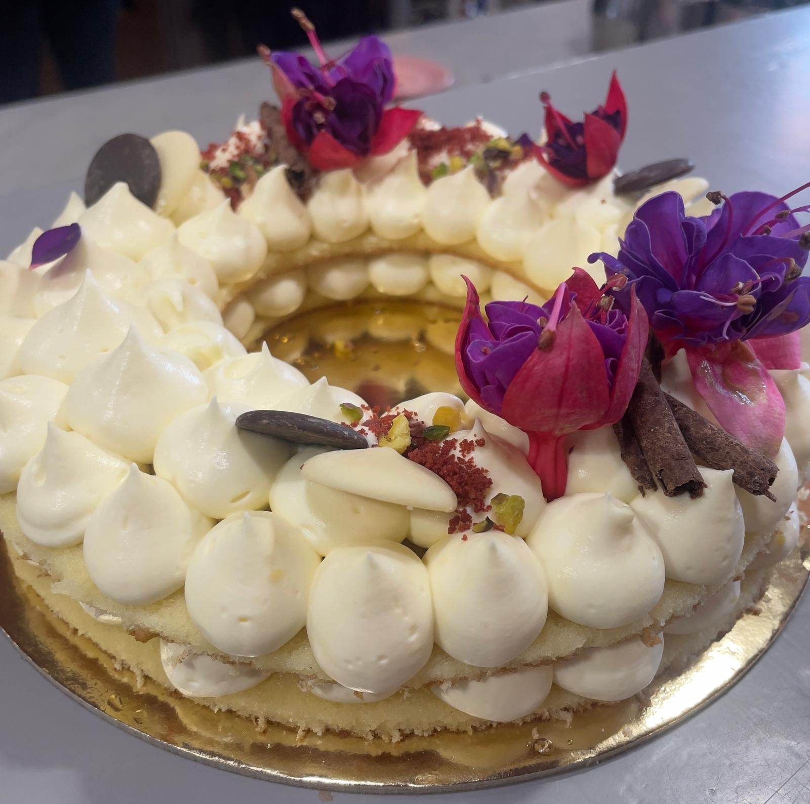 Torta a forma di ciambella con glassa bianca, decorata con fiori, cioccolato e pistacchi.