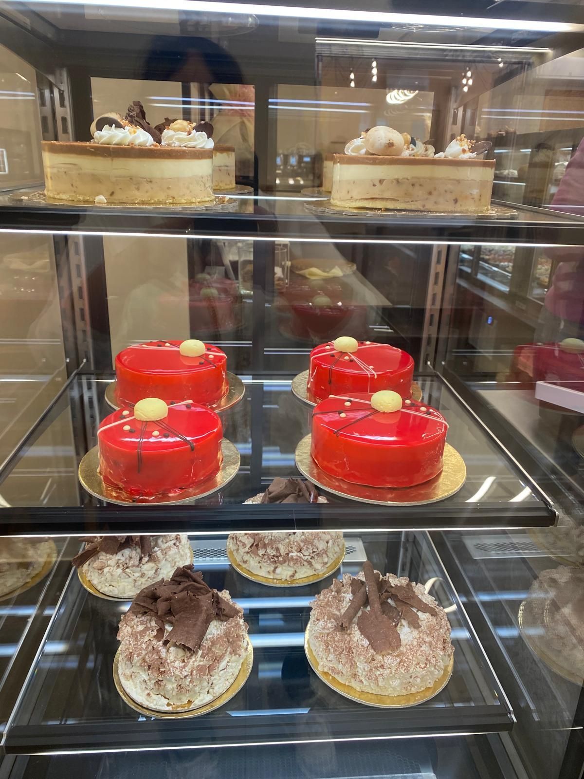 Torte in vetrina: glassate di rosso, al cioccolato e ricoperte di panna e frutta, in un ambiente da pasticceria.