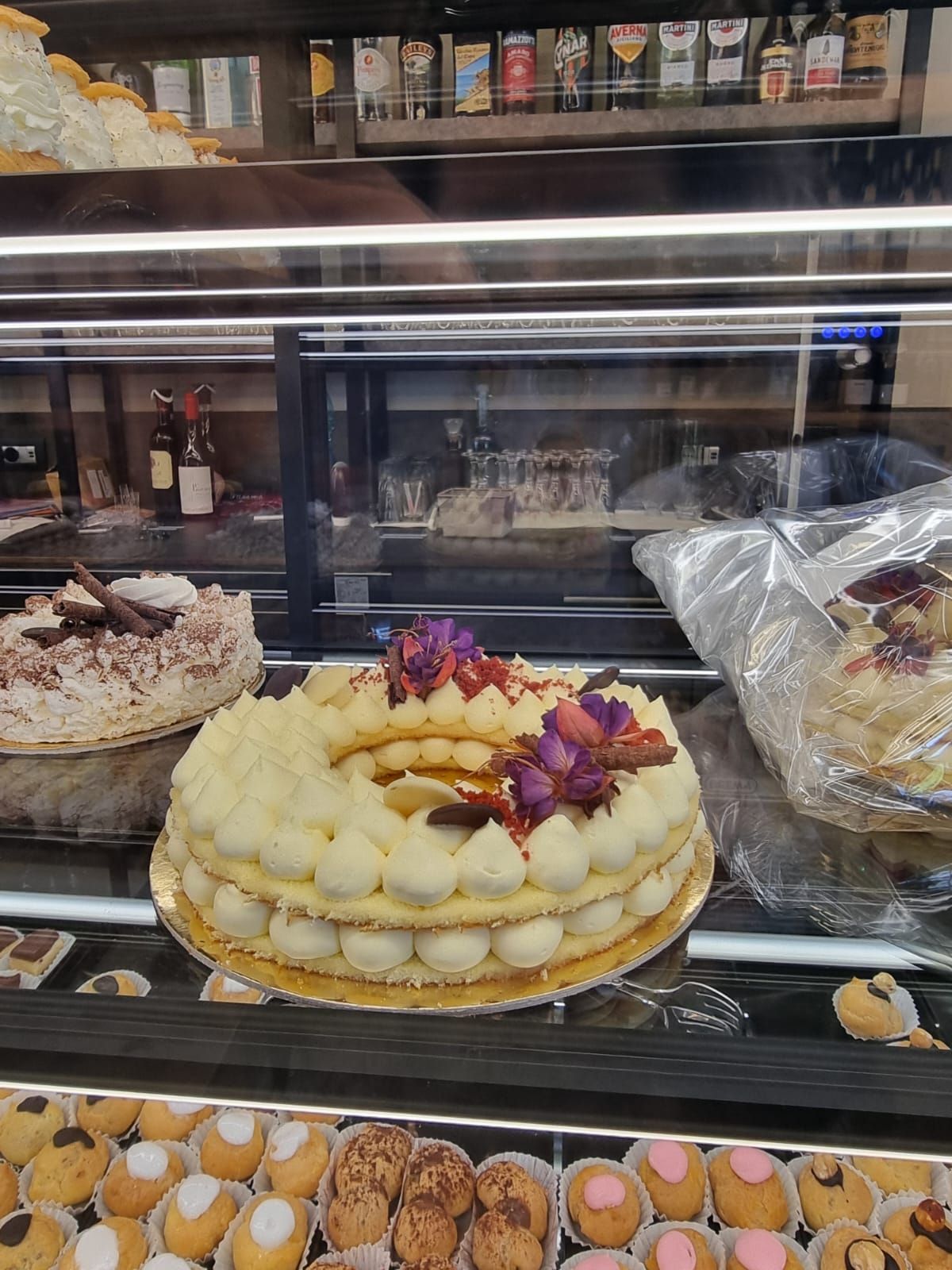 Vetrina di pasticceria con una ciambella decorata con fiori e panna. Altri dolci sono visibili.