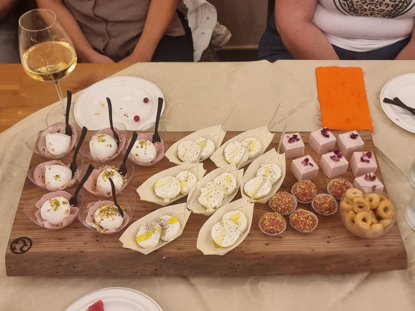 Tagliere di legno con antipasti, tra cui palline di formaggio, uova e mini tortine, con persone sedute attorno a un tavolo.