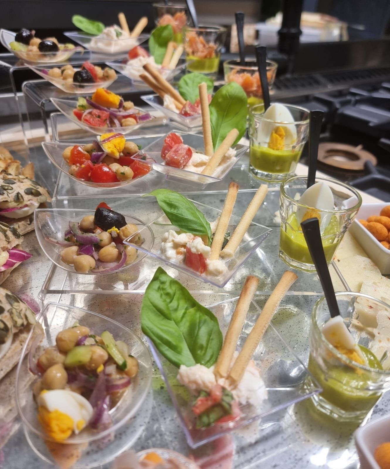 Buffet con vari antipasti di piccole dimensioni su vassoi trasparenti a più livelli; comprende insalate, creme spalmabili, uova sode e grissini.
