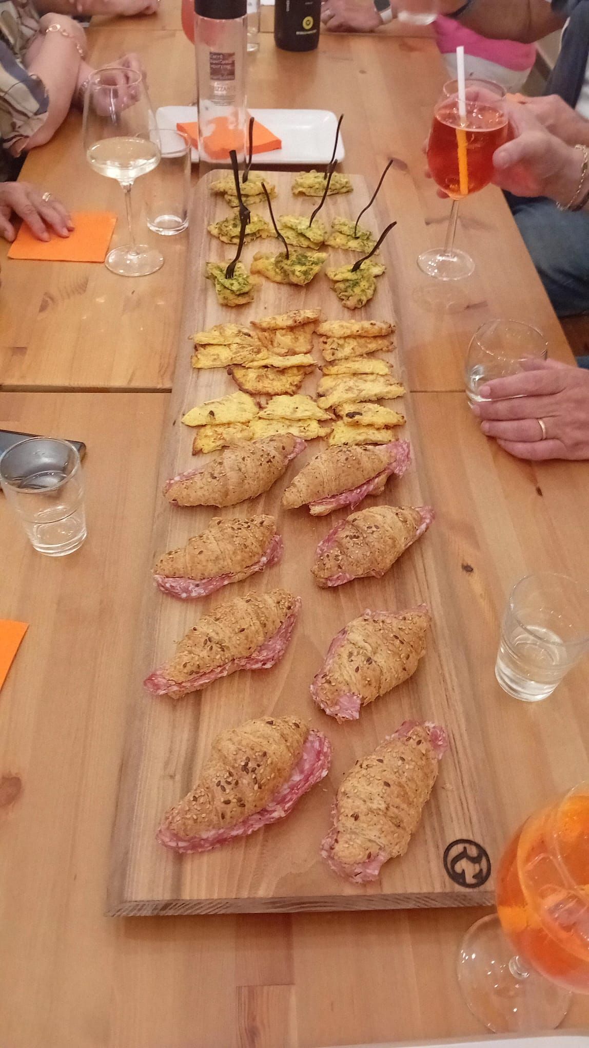 Tagliere di legno con vari stuzzichini, come croissant e cracker, su un tavolo con persone e bevande.