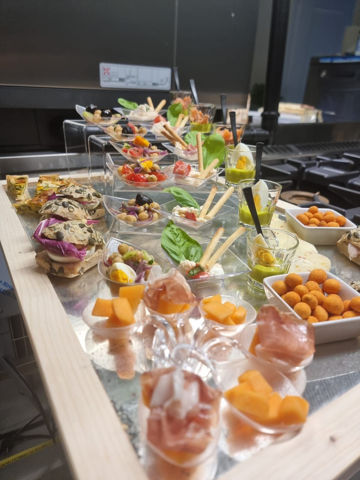 Antipasti assortiti esposti su una superficie metallica in una cucina. Include piccole porzioni di melone, prosciutto e altre prelibatezze.