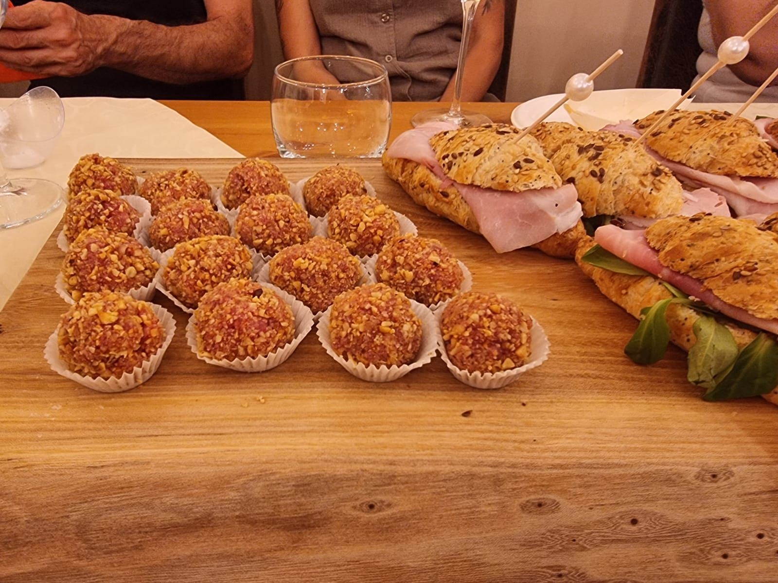 Un tagliere di legno con antipasti: polpette in bicchieri di carta e panini, con persone sullo sfondo.