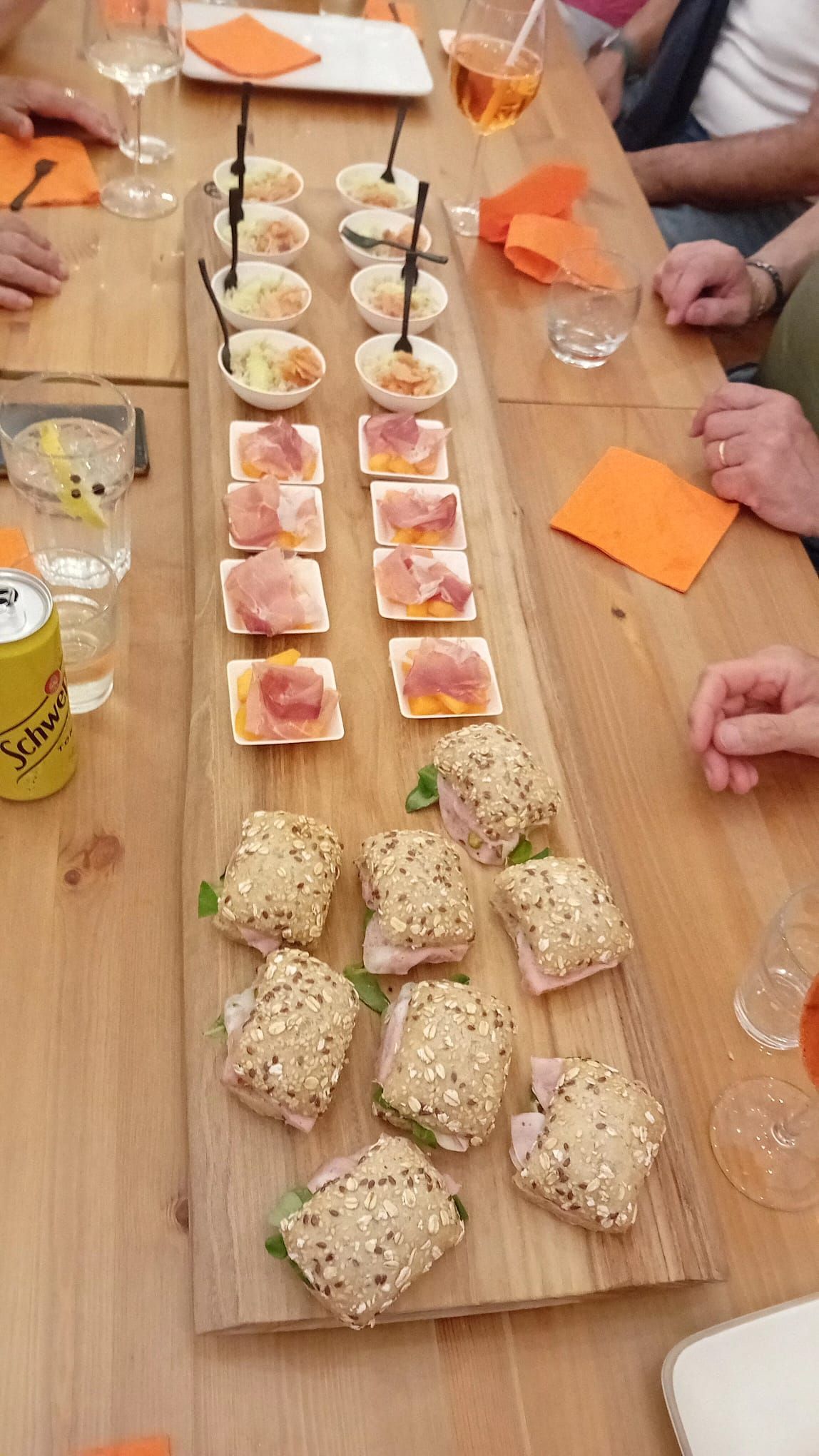 Tagliere di legno con antipasti su un tavolo: vari piccoli stuzzichini con prosciutto, panini e bevande.