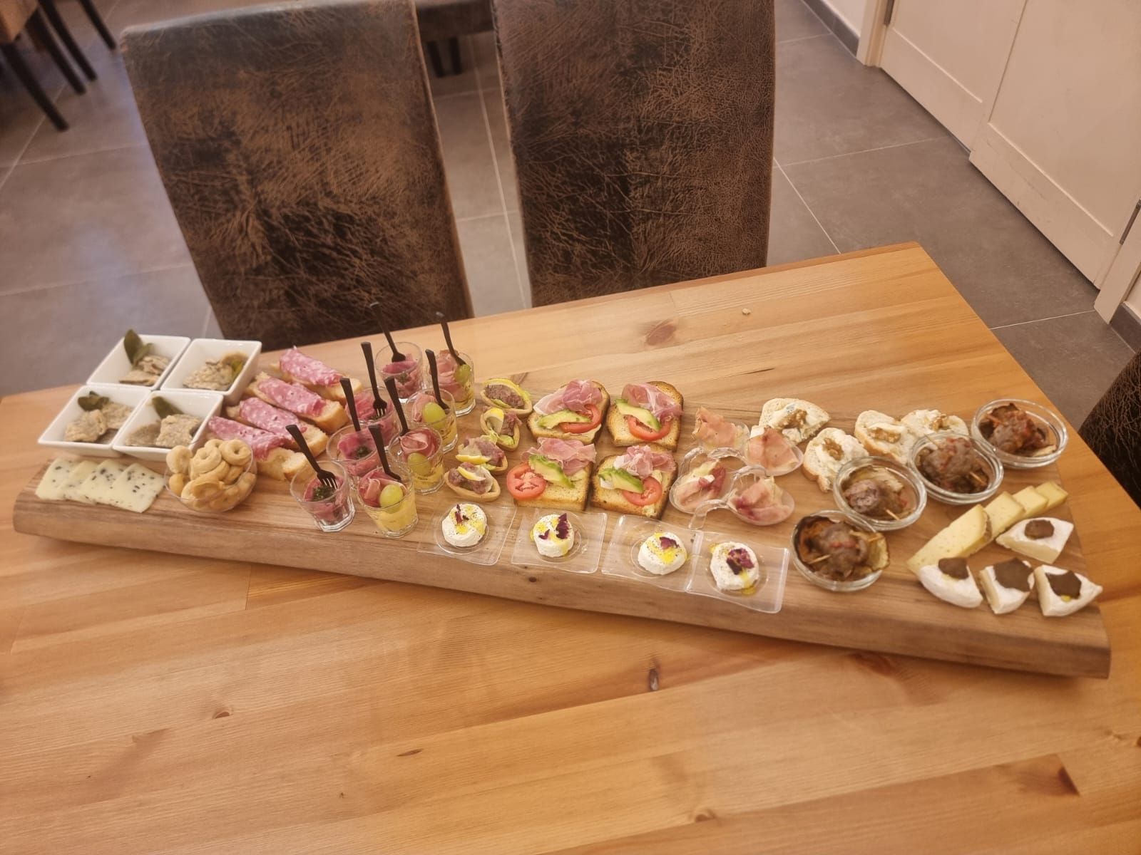 Vassoio di legno con antipasti: salumi, formaggi e piccola pasticceria. Disposto su un tavolo di legno, vicino a due sedie.