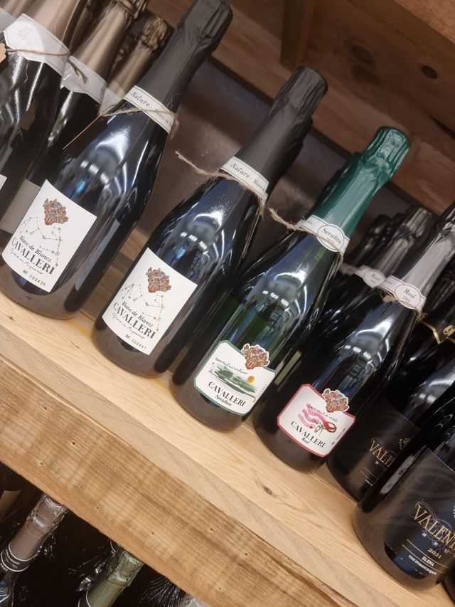 Bottiglie di spumante su uno scaffale di legno. Le bottiglie hanno tappi, etichette e spago neri. Alcune etichette sono marroni e bianche.