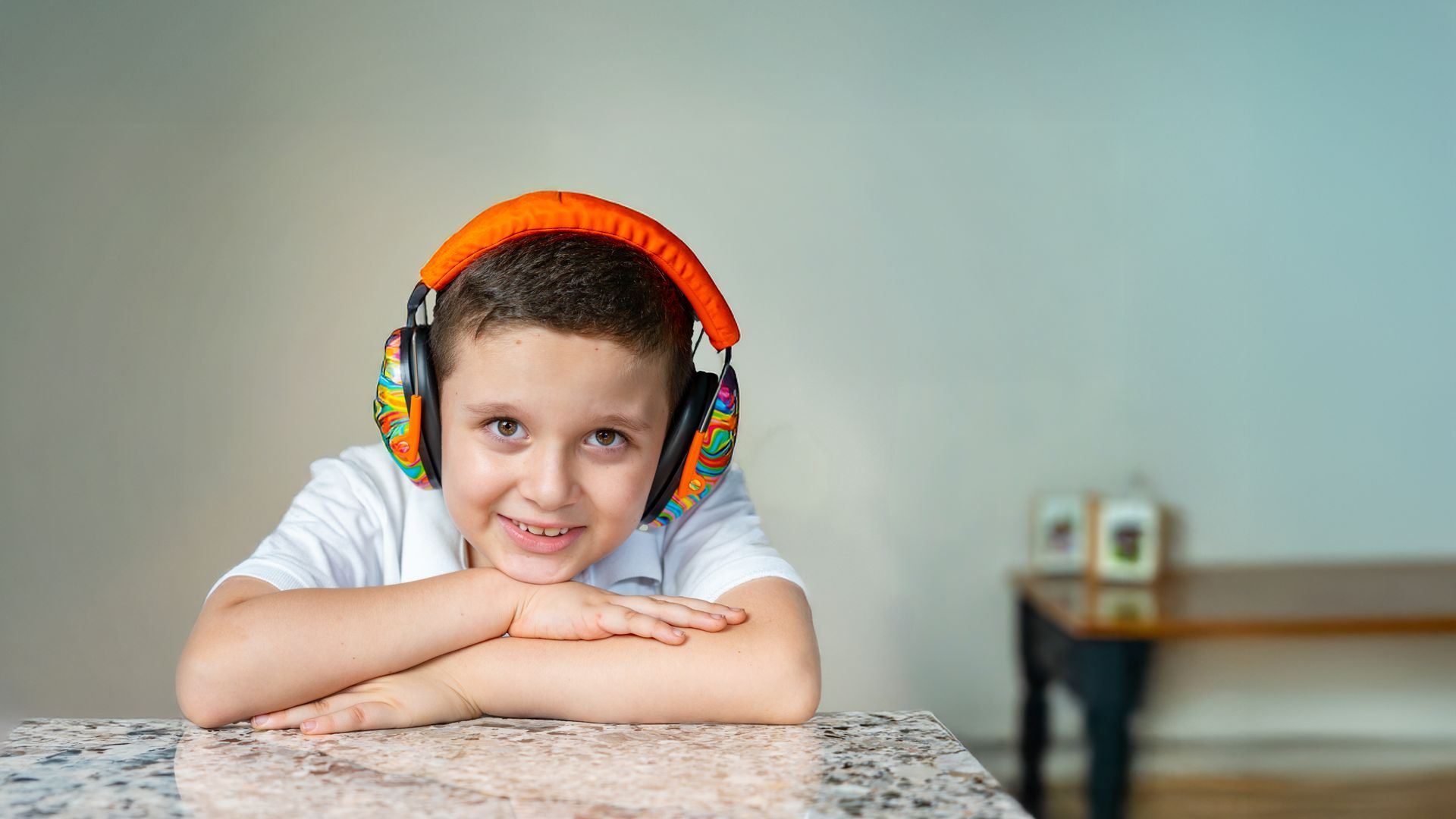 Child resting arms on a table, wearing orange headphones and smiling at the camera.